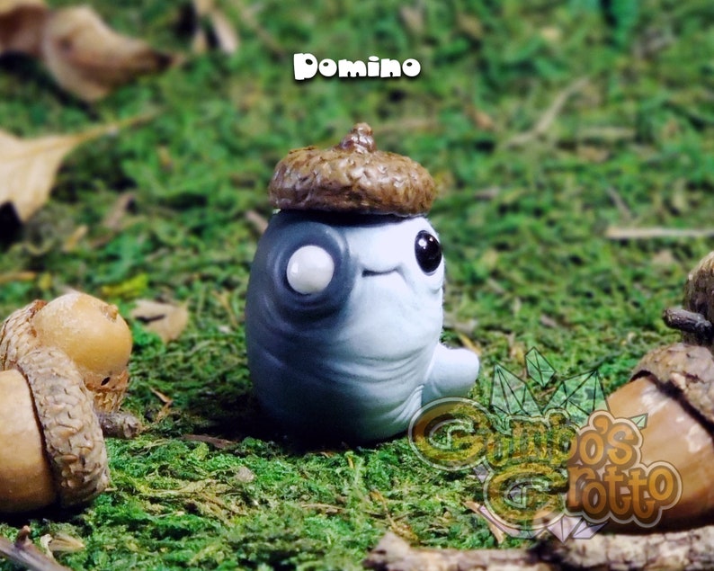 May include: A small, grey and white figurine with a brown acorn hat. The figurine has a single white eye and a small smile. The figurine is sitting on a bed of green moss.