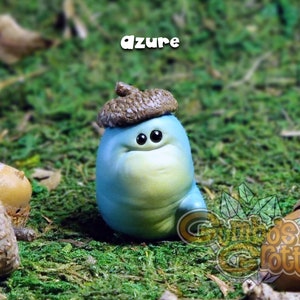 May include: A small, blue, hand-painted figurine of a worm wearing an acorn hat. The worm has a light blue body with a yellow belly and large, black eyes. The acorn hat is brown with a light brown cap. The figurine is sitting on a bed of green moss.