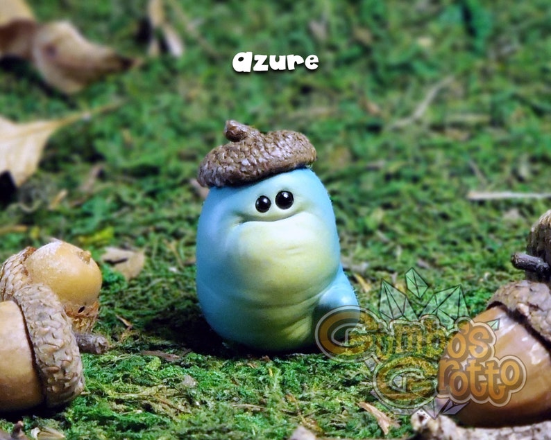 May include: A small, blue, hand-painted figurine of a worm wearing an acorn hat. The worm has a light blue body with a yellow belly and large, black eyes. The acorn hat is brown with a light brown cap. The figurine is sitting on a bed of green moss.