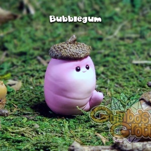 May include: A small pink and white figurine shaped like an acorn with a brown cap. The figurine has a smiling face and is sitting on a bed of green moss.