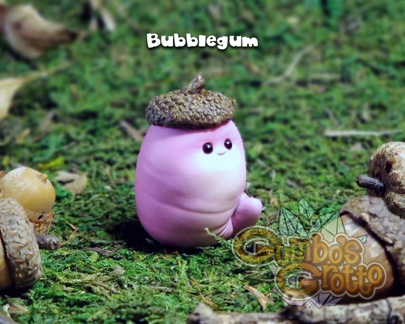 May include: A small pink and white figurine shaped like an acorn with a brown cap. The figurine has a smiling face and is sitting on a bed of green moss.