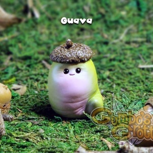 May include: A small, hand-painted figurine of a cartoon character with a pink body, green legs, and a brown acorn hat. The figurine is sitting on a bed of green moss. The text "Guava" is written in white letters above the figurine.