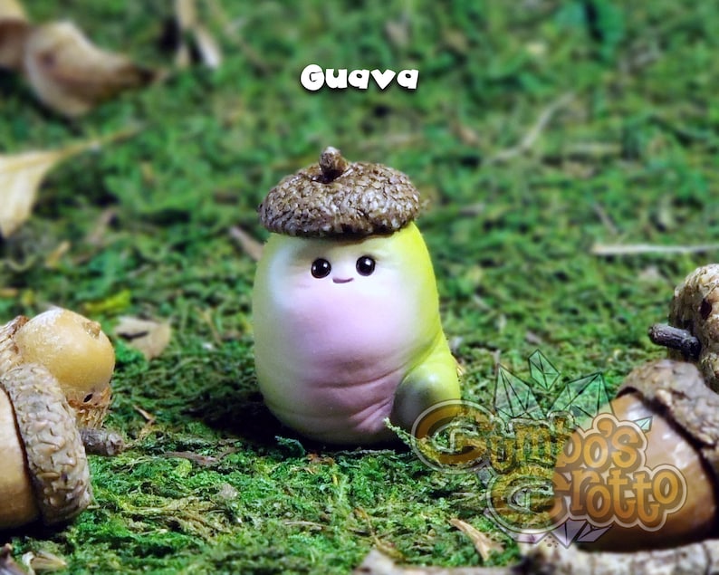 May include: A small, hand-painted figurine of a cartoon character with a pink body, green legs, and a brown acorn hat. The figurine is sitting on a bed of green moss. The text "Guava" is written in white letters above the figurine.