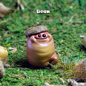 May include: A small, brown, ceramic figurine of a character with a large, round head and a smiling face. The figurine is wearing a brown acorn hat and is sitting on a bed of green moss. The figurine is labelled "Bean".