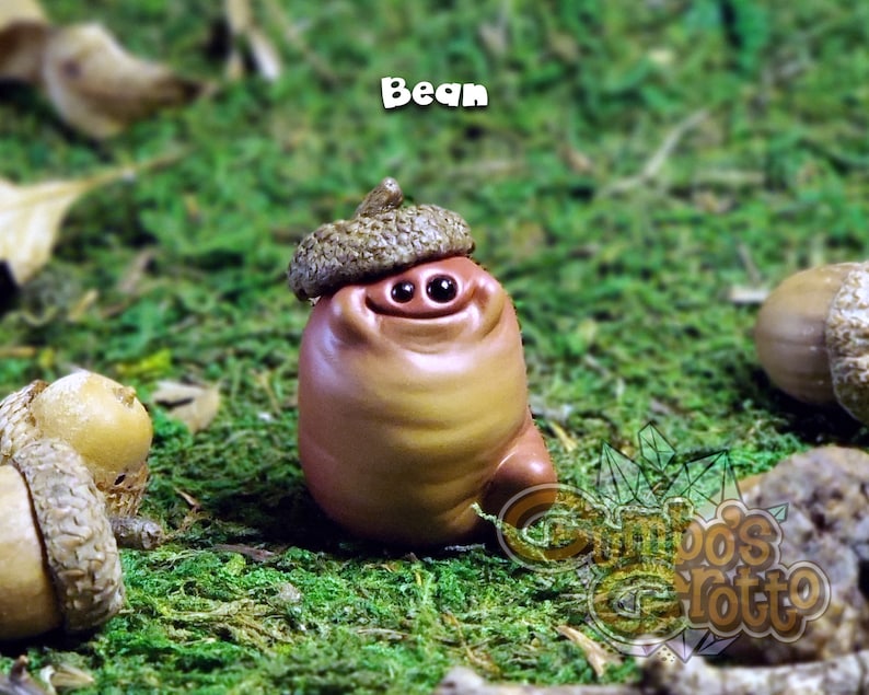 May include: A small, brown, ceramic figurine of a character with a large, round head and a smiling face. The figurine is wearing a brown acorn hat and is sitting on a bed of green moss. The figurine is labelled "Bean".