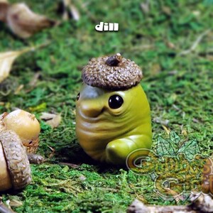 May include: A small green and brown figurine with a large acorn hat. The figurine has a smiling face and large eyes. The figurine is sitting on a bed of green moss.