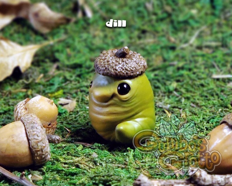 May include: A small green and brown figurine with a large acorn hat. The figurine has a smiling face and large eyes. The figurine is sitting on a bed of green moss.