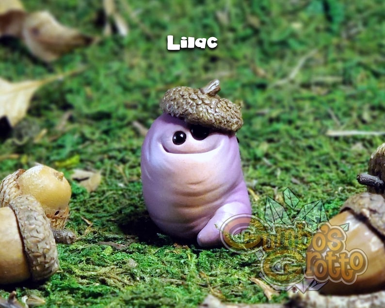 May include: A small, purple, clay figurine with a smiling face and a brown acorn hat. The figurine is sitting on a bed of green moss.