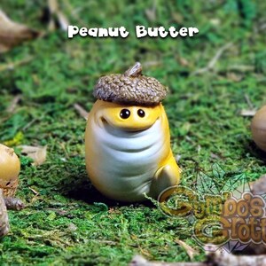 May include: A small, yellow figurine with a brown acorn hat. The figurine has a smiling face with large eyes and a white belly. The figurine is sitting on a bed of green moss. The text "Peanut Butter" is written above the figurine.