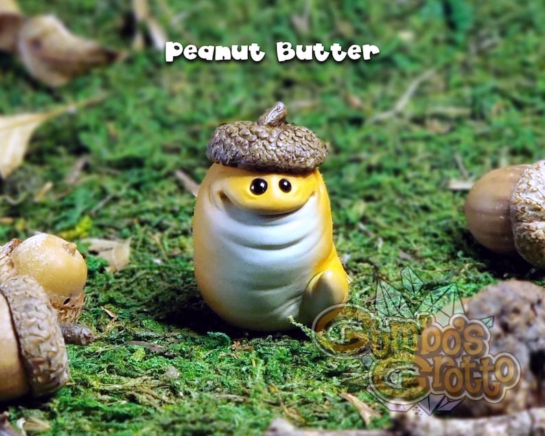 May include: A small, yellow figurine with a brown acorn hat. The figurine has a smiling face with large eyes and a white belly. The figurine is sitting on a bed of green moss. The text "Peanut Butter" is written above the figurine.