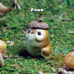May include: A small, brown, ceramic figurine of a fawn wearing an acorn hat. The figurine is sitting on a bed of green moss.