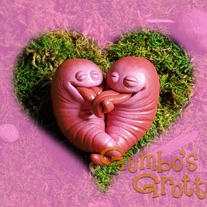 May include: Two pink clay figures of a creature with closed eyes and a smile, cuddling inside a heart-shaped moss bed. The figures are in a heart-shaped bed of moss.