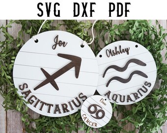 May include: Two white wooden round signs with brown wood zodiac symbols and names. The first sign has a brown Sagittarius symbol and the name "Joe". The second sign has a brown Aquarius symbol and the name "Ashley". A smaller white wooden round sign with a brown Cancer symbol and the name "Dean" is in the foreground.