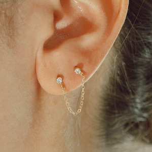 Puede incluir: Un par de pendientes de oro con pequeños diamantes, conectados por una delicada cadena de oro.