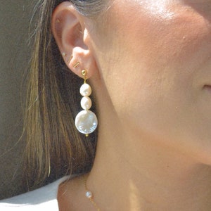 May include: A woman wearing a white top and a gold necklace with pearl accents. She is wearing a pair of gold earrings with a large pearl and smaller pearls hanging from it.