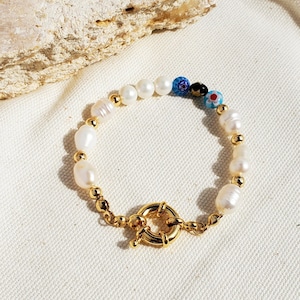 Puede incluir: Un brazalete dorado con perlas blancas y cuentas de vidrio de colores. El brazalete tiene un cierre de palanca.