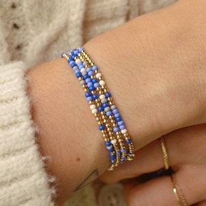 Puede incluir: Pulsera de cuentas azules, blancas y doradas en la muñeca. La pulsera está compuesta por múltiples hilos. La persona lleva un cárdigan de punto trenzado color crema. También se ve un anillo dorado.