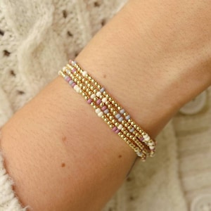 Puede incluir: Un conjunto de cuatro pulseras delicadas con pequeñas cuentas doradas y cuentas de colores pastel. Las pulseras están superpuestas en la muñeca de una persona.