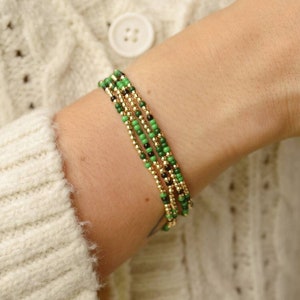 Puede incluir: Un brazalete de capas con cuentas verdes y doradas en una muñeca.