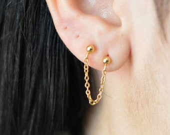 Puede incluir: Primer plano de una oreja con dos pendientes dorados. Cada pendiente presenta un pequeño perno de bola conectado por una delicada cadena a otro perno de bola. Los pendientes son de color dorado brillante.