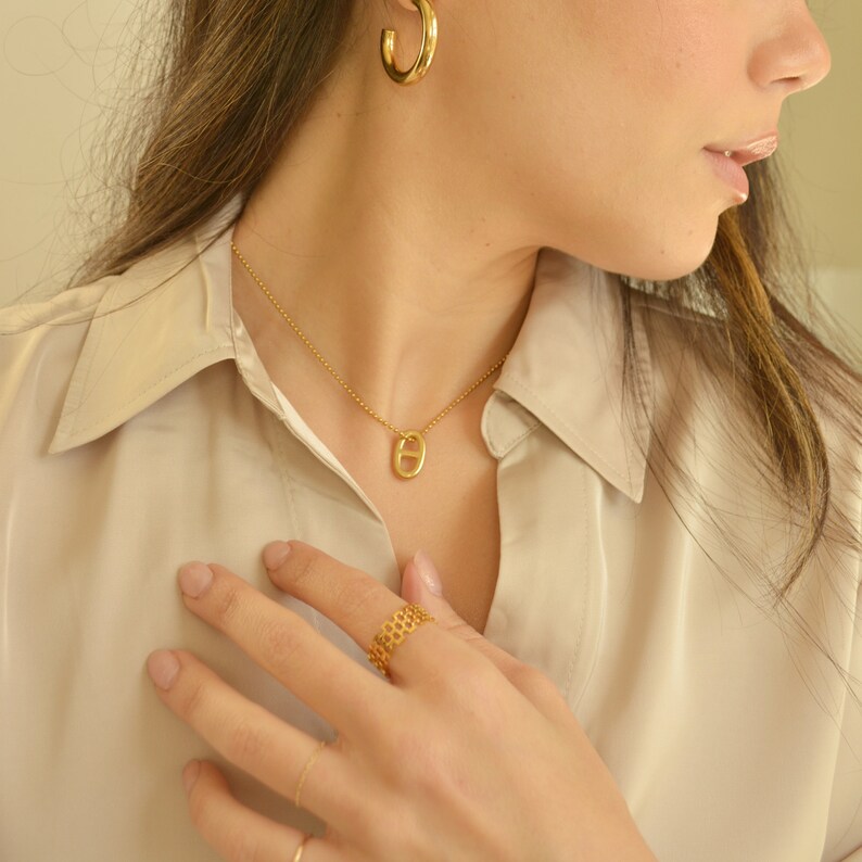 May include: A gold-toned jewelry set featuring a necklace with a pendant, a ring, and hoop earrings. The necklace has a delicate chain with a small, oval-shaped pendant. The ring has a honeycomb design. The hoop earrings are simple and classic.
