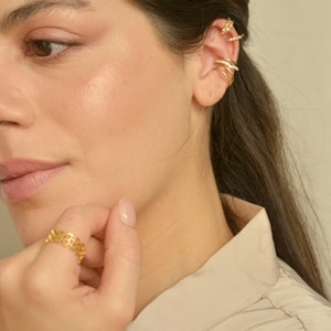 May include: A close-up of a person wearing gold jewelry. The jewelry includes multiple ear cuffs and a ring. The ear cuffs are adorned with small, sparkling stones. The ring has a geometric design.