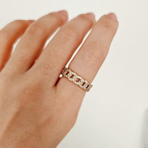 Puede incluir: Un anillo dorado con eslabones de cadena y piedras claras y brillantes engastadas en los eslabones. El anillo está en un dedo, sobre un fondo blanco. El diseño del anillo es un patrón continuo de eslabones entrelazados.