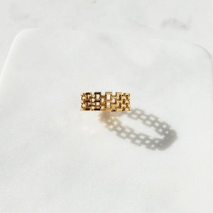May include: A gold-colored ring with a rectangular chain-link design. The ring sits on a white surface, casting a shadow. The jewelry piece is a band style, with a repeating pattern of open rectangles.