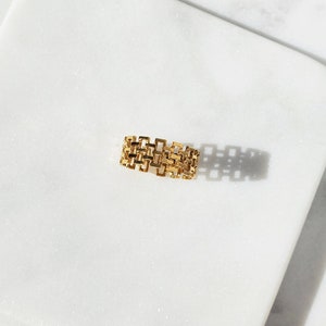 May include: A gold-colored ring with a geometric design. The ring features a series of interconnected rectangular shapes, creating an open, textured surface. The ring is set against a white marble background, with shadows adding depth.