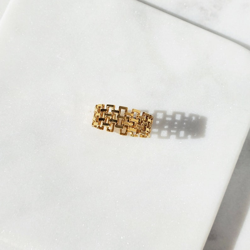 May include: A gold-colored ring with a geometric design. The ring features a series of interconnected rectangular shapes, creating an open, textured surface. The ring is set against a white marble background, with shadows adding depth.