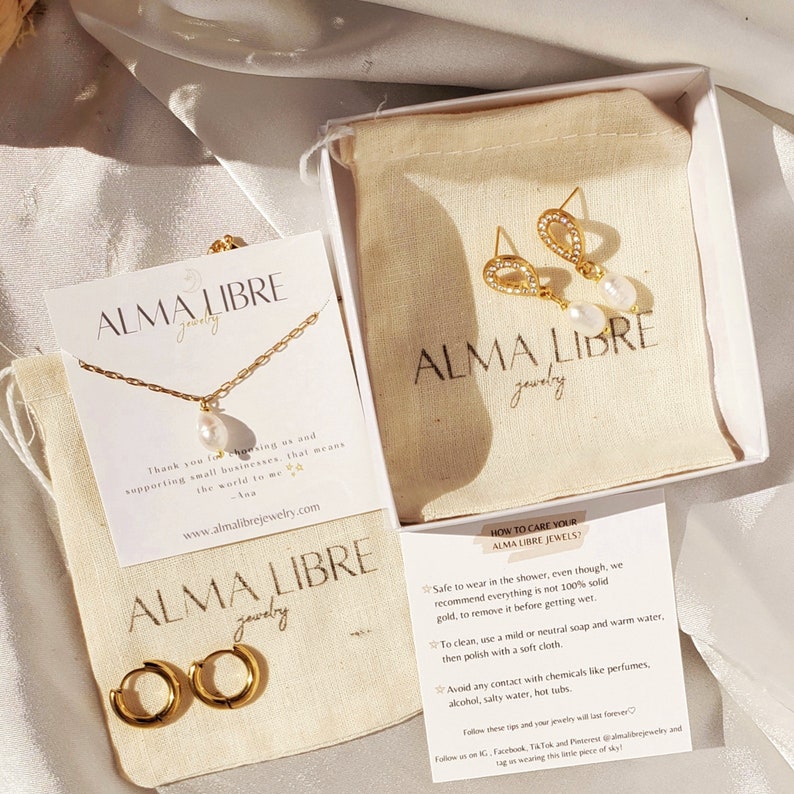 May include: A gold-toned jewelry set featuring a necklace with a pearl pendant, hoop earrings, and teardrop earrings with pearls. The set includes cards with care instructions and the brand name "ALMA LIBRE jewelry". The items are displayed on a white surface.