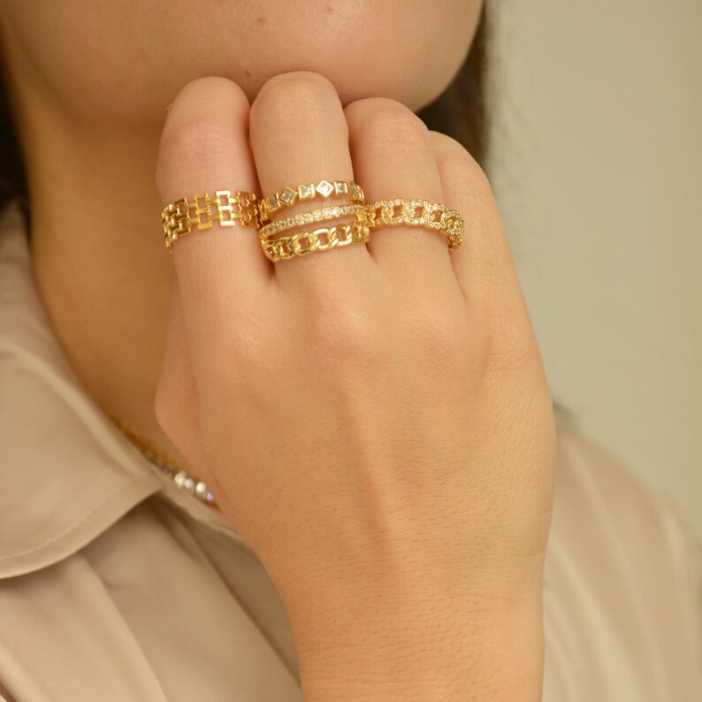 May include: A hand wearing four gold rings. The rings feature various designs, including a chain link, a rectangular pattern, and a row of small diamonds. The rings are displayed against a neutral background.