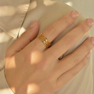 May include: A gold-colored ring with a geometric, openwork design. The ring features a series of connected rectangular shapes, creating a mesh-like pattern. The ring is worn on a finger, showcasing its intricate details and reflective surface.