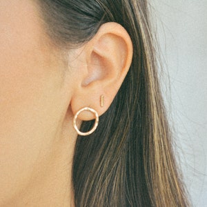 Puede incluir: Primer plano de la oreja de una persona con dos pendientes de oro. El pendiente del lóbulo inferior es un círculo martillado, y el pendiente del lóbulo superior es una barra fina y recta.