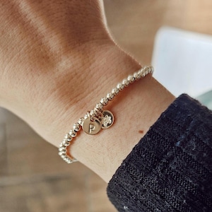 Puede incluir: Pulsera de cuentas plateadas con dos colgantes. Un colgante es un pequeño disco redondo con la letra "P" grabada. El otro colgante es una pequeña flor. La pulsera se lleva en la muñeca.