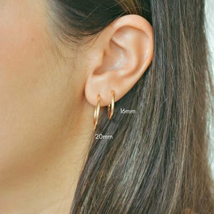 Puede incluir: Primer plano de dos pendientes de aro dorados, uno de 16 mm y otro de 20 mm de diámetro, en una oreja. Los pendientes son simples, circulares y brillantes. El fondo muestra la piel y el cabello.