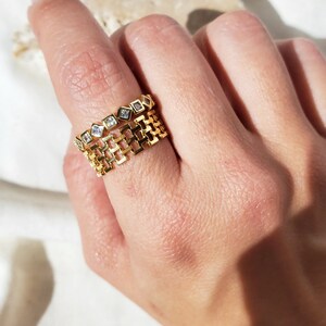 May include: Two gold rings are displayed on a finger. The top ring features a series of small, square, clear gemstones. The bottom ring has a chain-link design. The rings are made of gold-colored metal and are set against a soft, neutral background.