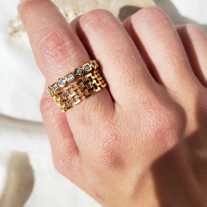 May include: Two gold rings are displayed on a finger. The top ring features a series of small, square, clear gemstones. The bottom ring has a chain-link design. The rings are made of gold-colored metal and are set against a soft, neutral background.