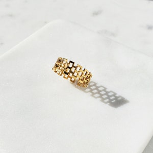 May include: A gold-colored ring with a geometric, openwork design. The ring features a series of interconnected rectangular shapes, creating a lattice-like pattern. The ring is displayed on a white surface, casting a shadow.
