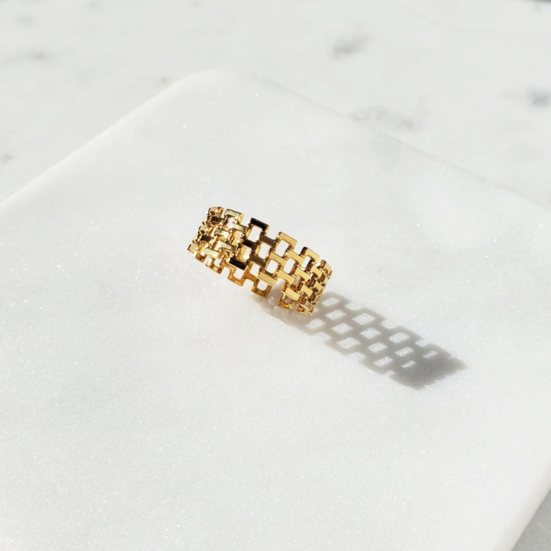May include: A gold-colored ring with a geometric, openwork design. The ring features a series of interconnected rectangular shapes, creating a lattice-like pattern. The ring is displayed on a white surface, casting a shadow.