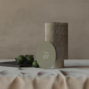 May include: A light green round table number card with the text "Table 07" on a white background. The card is sitting on top of a textured, light brown cylinder. There are green limes on a dark brown rectangular surface in the foreground.