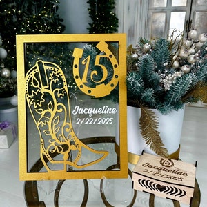 May include: Gold-colored frame with a cutout of a cowboy boot and horseshoe with the number 15 inside. The frame has the name Jacqueline and the date 2/22/2025. A small wooden box with the same design and text is in the foreground.
