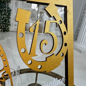 May include: A gold-painted wooden sign with the number "15" inside a horseshoe shape. The sign is mounted on a wooden frame.