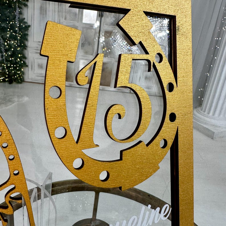 May include: A gold-painted wooden sign with the number "15" inside a horseshoe shape. The sign is mounted on a wooden frame.