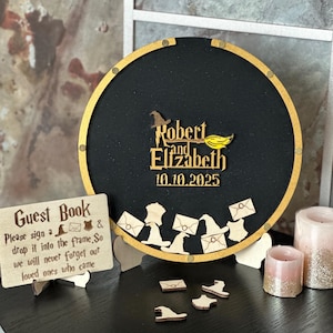 May include: A black circular wedding guest book with a gold frame, featuring the names "Robert and Elizabeth" and the date "10.10.2025". Includes wooden cutouts of hats, envelopes, and a sign that reads "Guest Book".