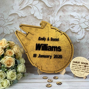 Puede incluir: Un letrero de madera dorado con forma de Halcón Milenario de Star Wars. El letrero tiene los nombres "Emily & Daniel Williams" y la fecha "18 de enero de 2020" impresos en él. También se ve en la imagen un letrero de madera redondo más pequeño con el texto "GUEST BOOK".