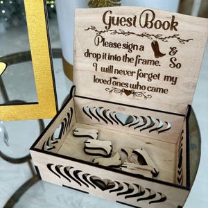May include: Wooden guest book box with a cutout design of hearts. The lid has a sign that reads "Guest Book Please sign a & drop it into the frame. So I will never forget my loved ones who came".