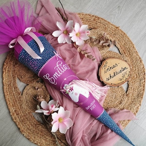 Könnte beinhalten: Eine handgefertigte lila und blaue Schultüte mit einem Einhorn-Design und dem Namen "Emilia" in Schreibschrift. Die Tüte ist mit rosa Tüll, Bändern und einem Holzschild verziert. Die Tüte ist etwa 71 cm lang.