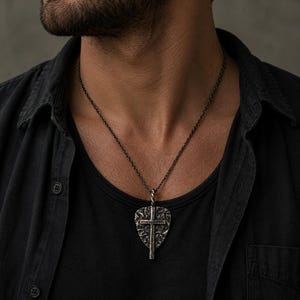 May include: A silver-colored pendant necklace featuring a guitar pick shape with a cross design. The pendant has a textured surface and hangs from a dark chain. The necklace is worn over a black shirt.