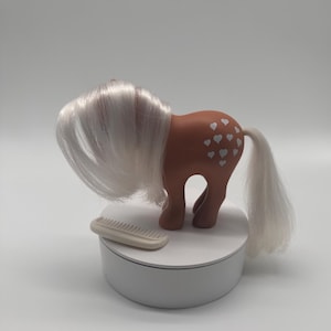 May include: A vintage toy pony with a brown body and white hair and tail. The pony has a series of white heart-shaped markings on its side. A small, off-white comb rests beside the pony on a white platform.